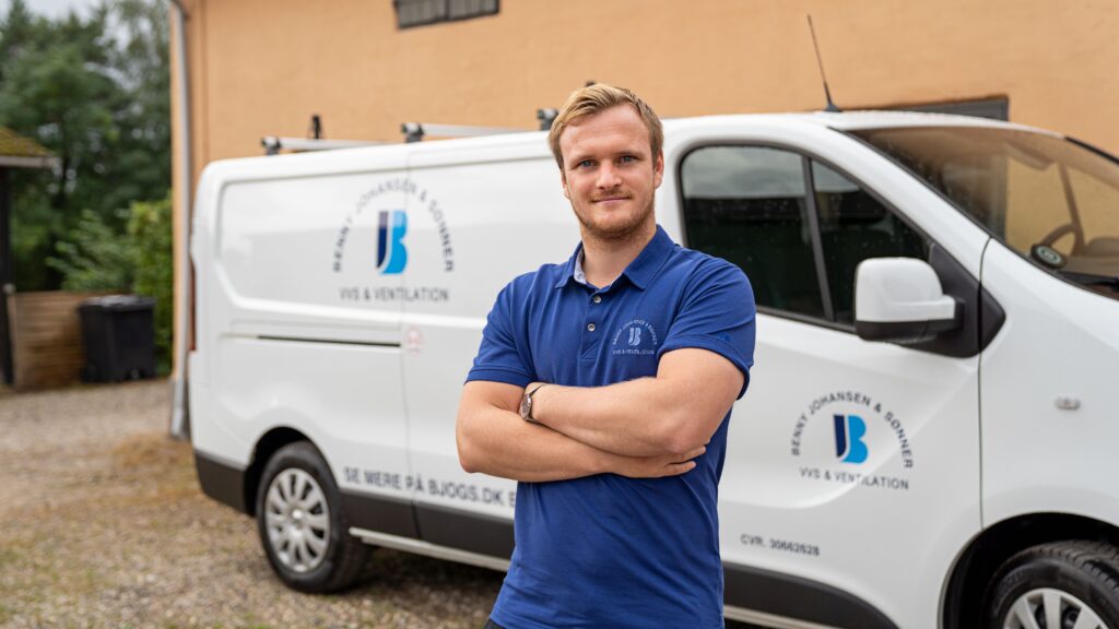 VVS’er Jonas Johansen der står i en blå t-shirt og kigger i kameraet med korslagte arme foran sin hvide firma varevogn